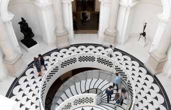 View from above looking down at the Tate Britain Rotunda terrazzo. A circular staircase leads down from a round gallery with statues on alcoves. There are a few visitors looking at the statues and walking down the stairs.