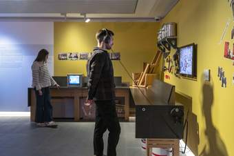 A photograph of a visitor standing by a display wearing headphones. There is another visitor in the background looking at a tv screen displayed on a work bench.